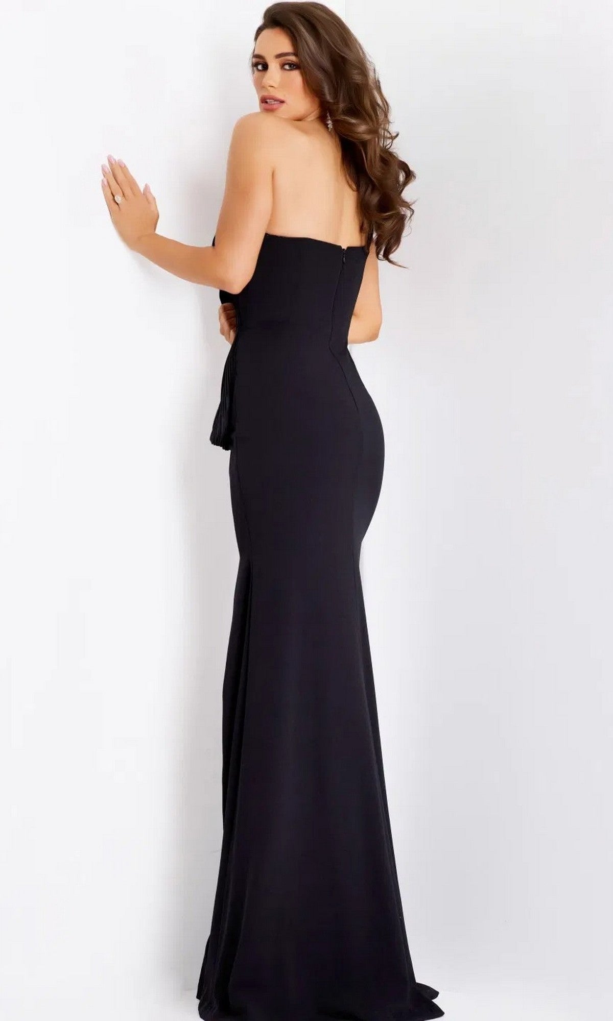 A woman with long wavy hair wears the Long Prom Dress: Jovani 43970 and stands facing a white wall, looking over her shoulder.